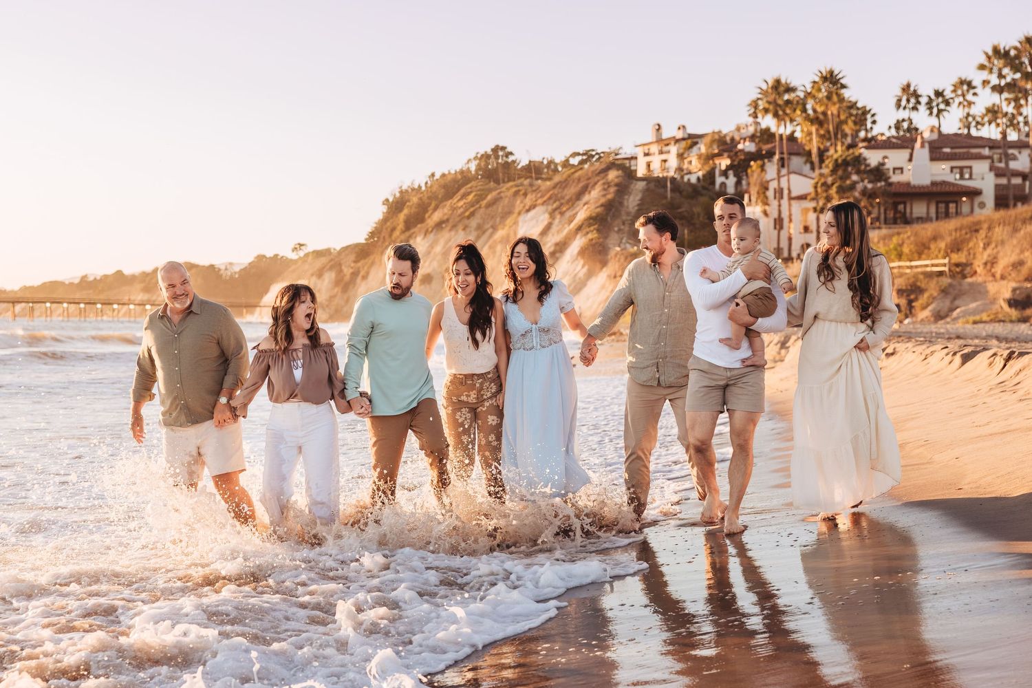 Large family walking along sunny beach shoreline at golden hour, wearing coordinated white and neutral outfits.