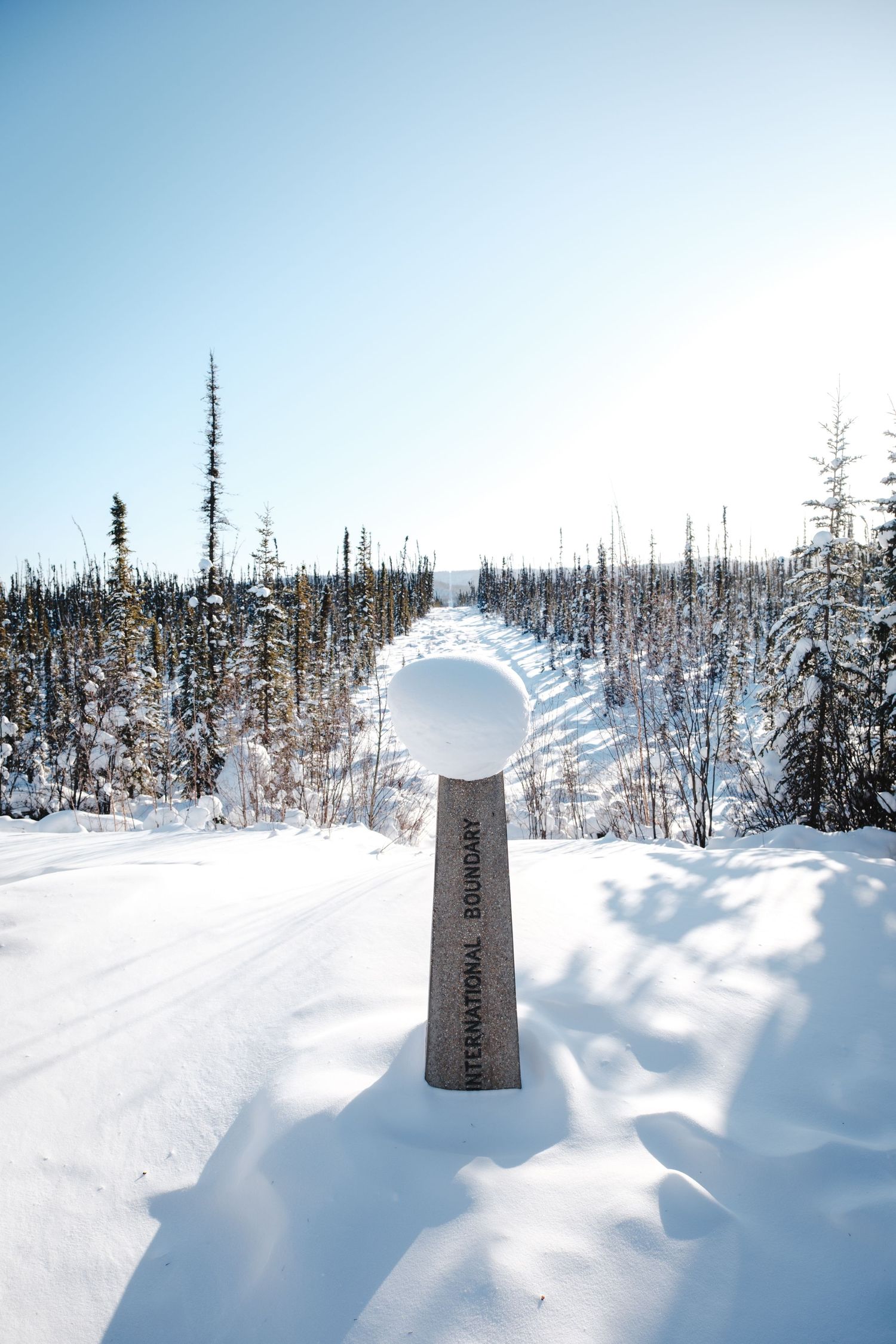 Snow-covered international boundary marker post standing in deep winter snow surrounded by frost-laden spruce trees.