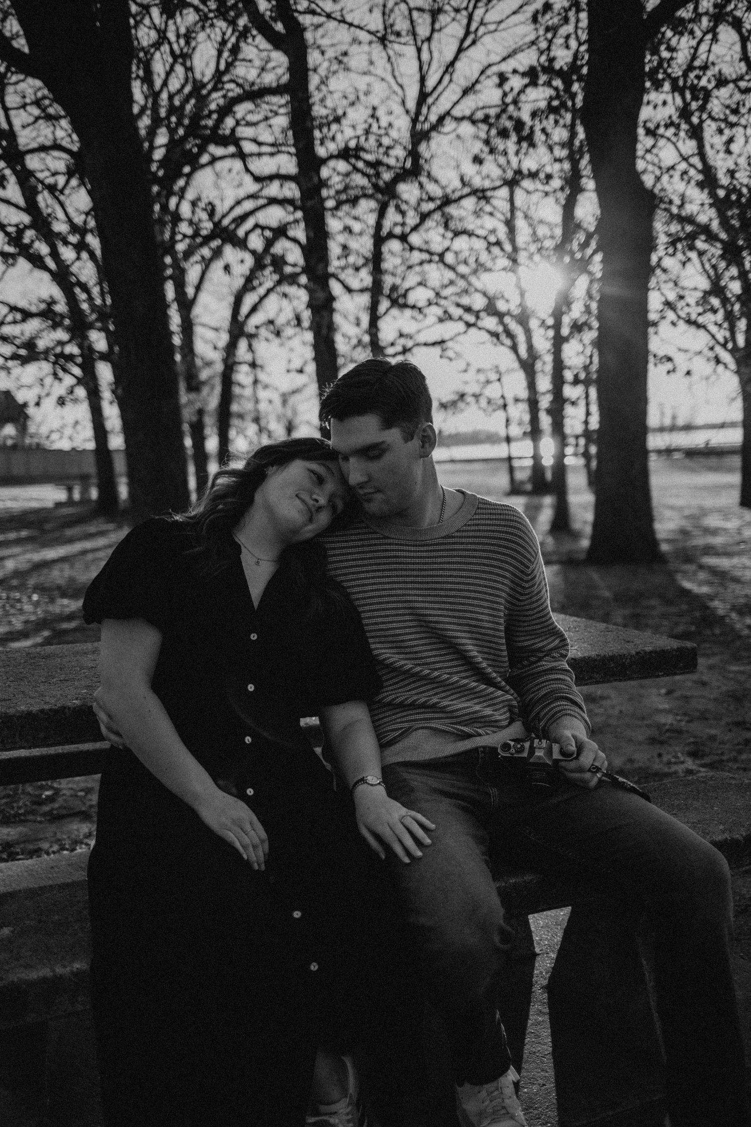 A couple sits closely together on a bench in a park at sunset in black and white.