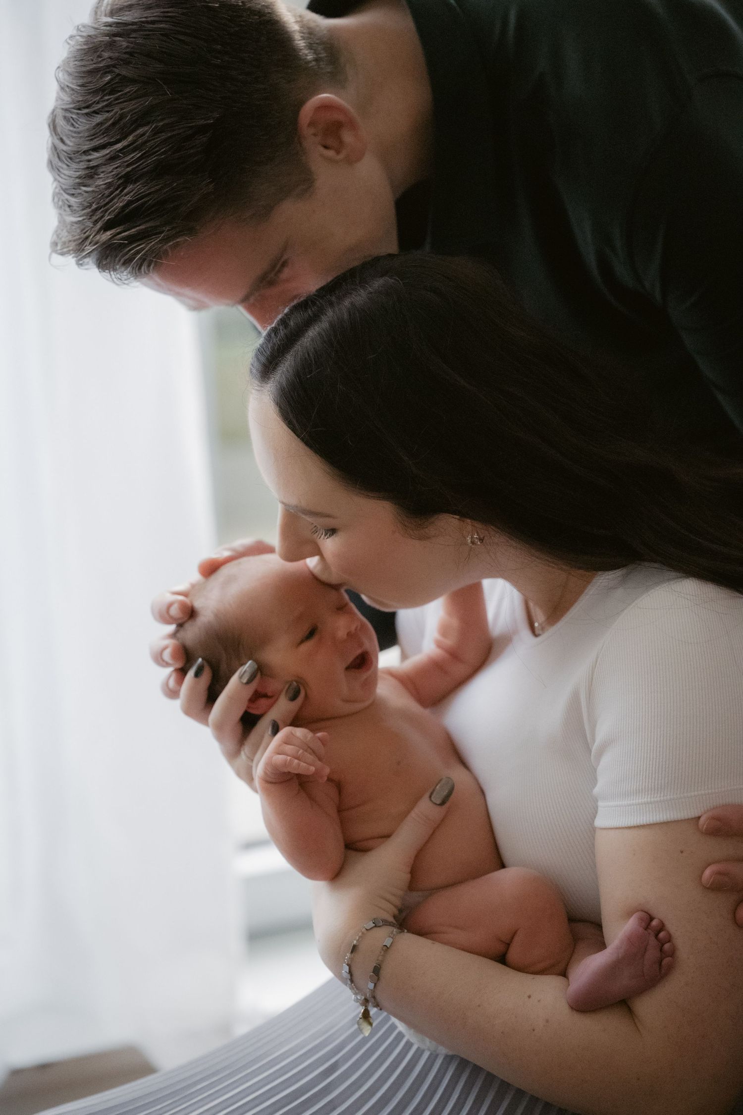New mom kissing newborn baby’s head with dad looking on, lifestyle newborn session Vancouver BC