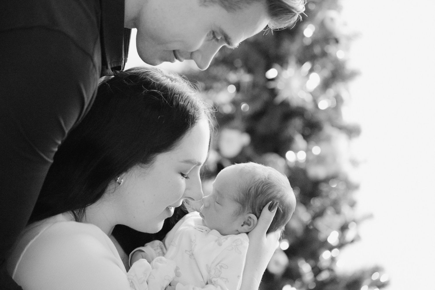 Black and white photo of parents cuddling their newborn baby with their Christmas tree in the background during in-home photo session