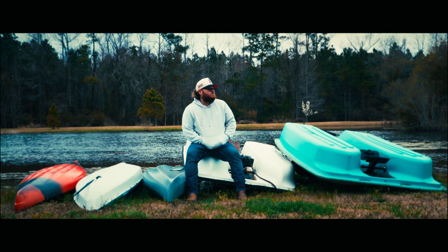 Man sitting on colorful kayaks by a calm lake surrounded by bare winter trees.