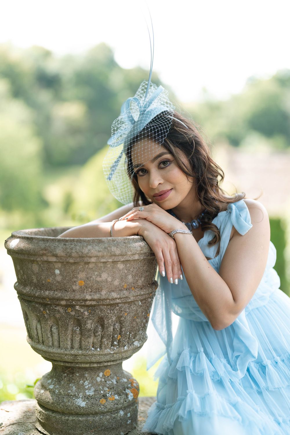 Woman in light blue tulle dress and fascinator hat leaning on stone urn in garden setting.