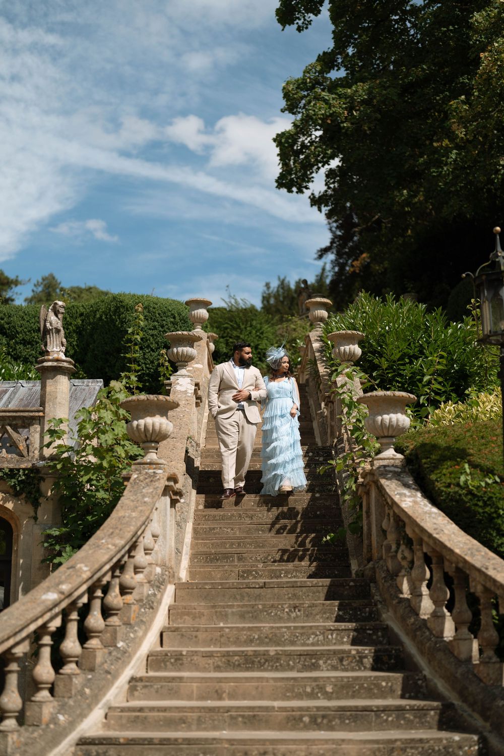 Elegant couple in formal attire descending grand stone staircase in lush garden setting under blue sky.