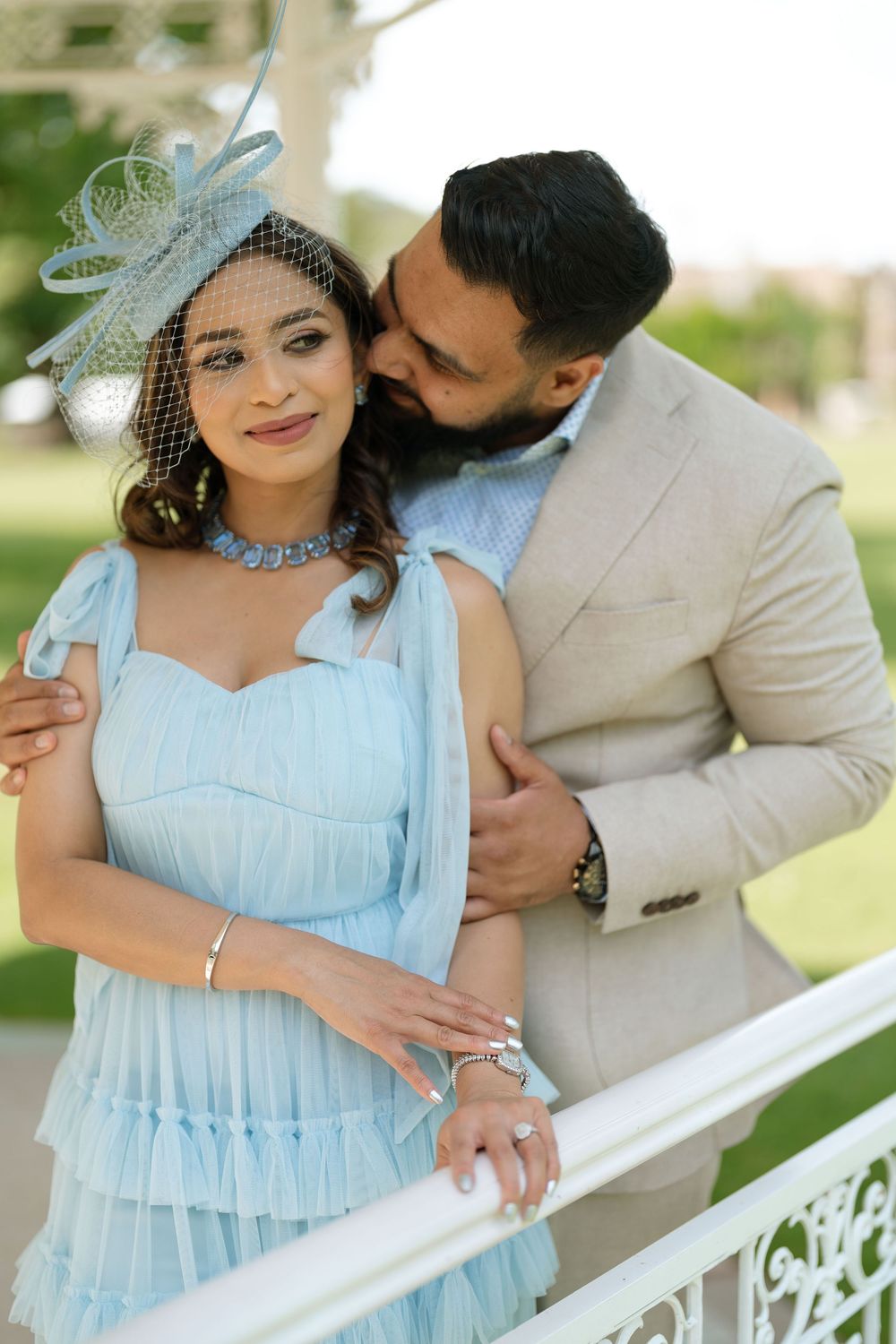 Elegant couple posing outdoors, woman in light blue dress with fascinator, man in beige suit kissing her cheek.