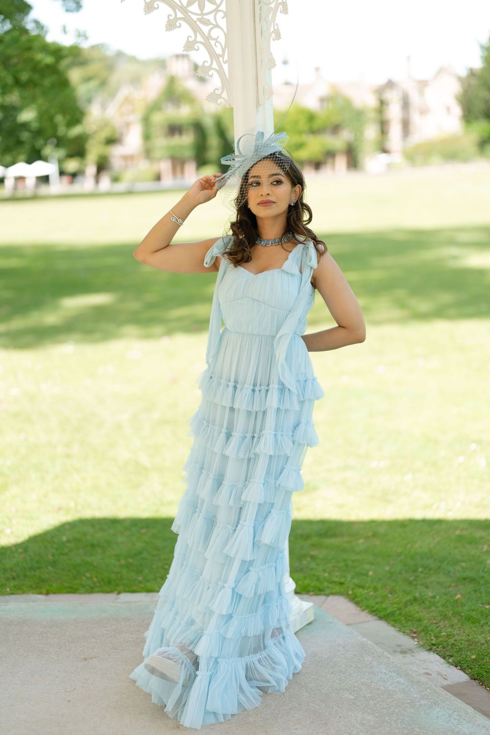 Woman in light blue tiered ruffle maxi dress and fascinator hat posing outdoors in a green park.