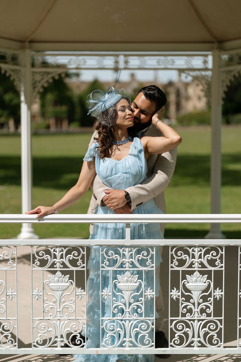 Elegant couple embracing on ornate white gazebo railing in formal attire, woman in blue dress with fascinator hat.