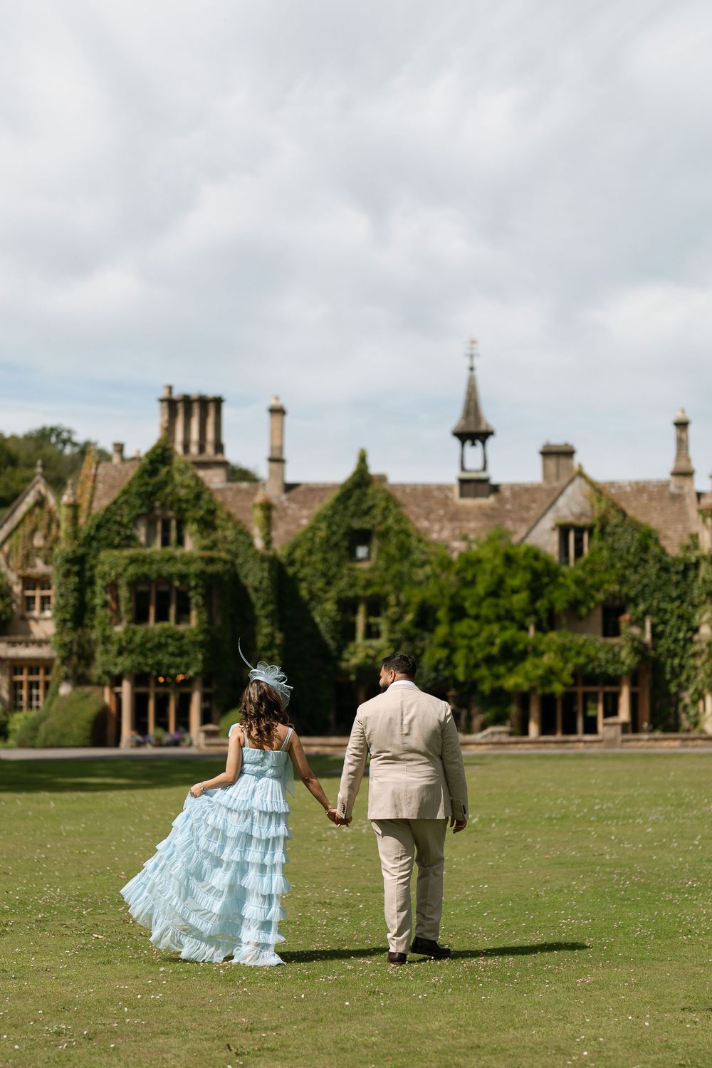 Couple in blue ruffled gown and beige suit holding hands walking toward ivy-covered English manor on green lawn.