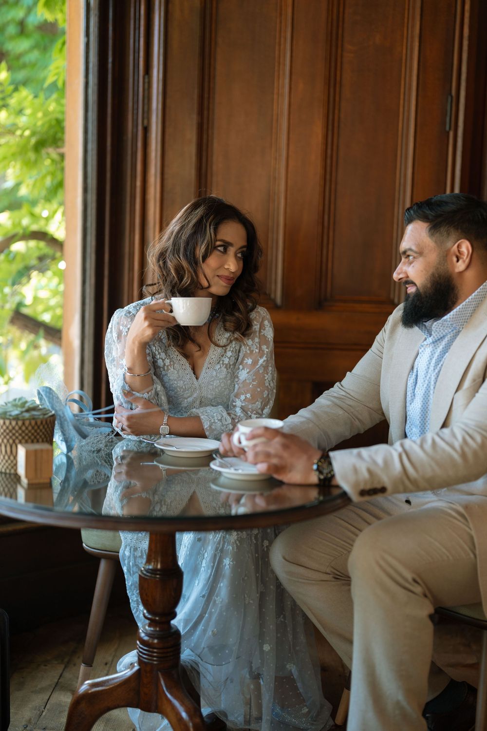 Elegant couple sharing coffee at a glass table in a luxurious wooden interior, woman in blue lace dress smiling warmly.
