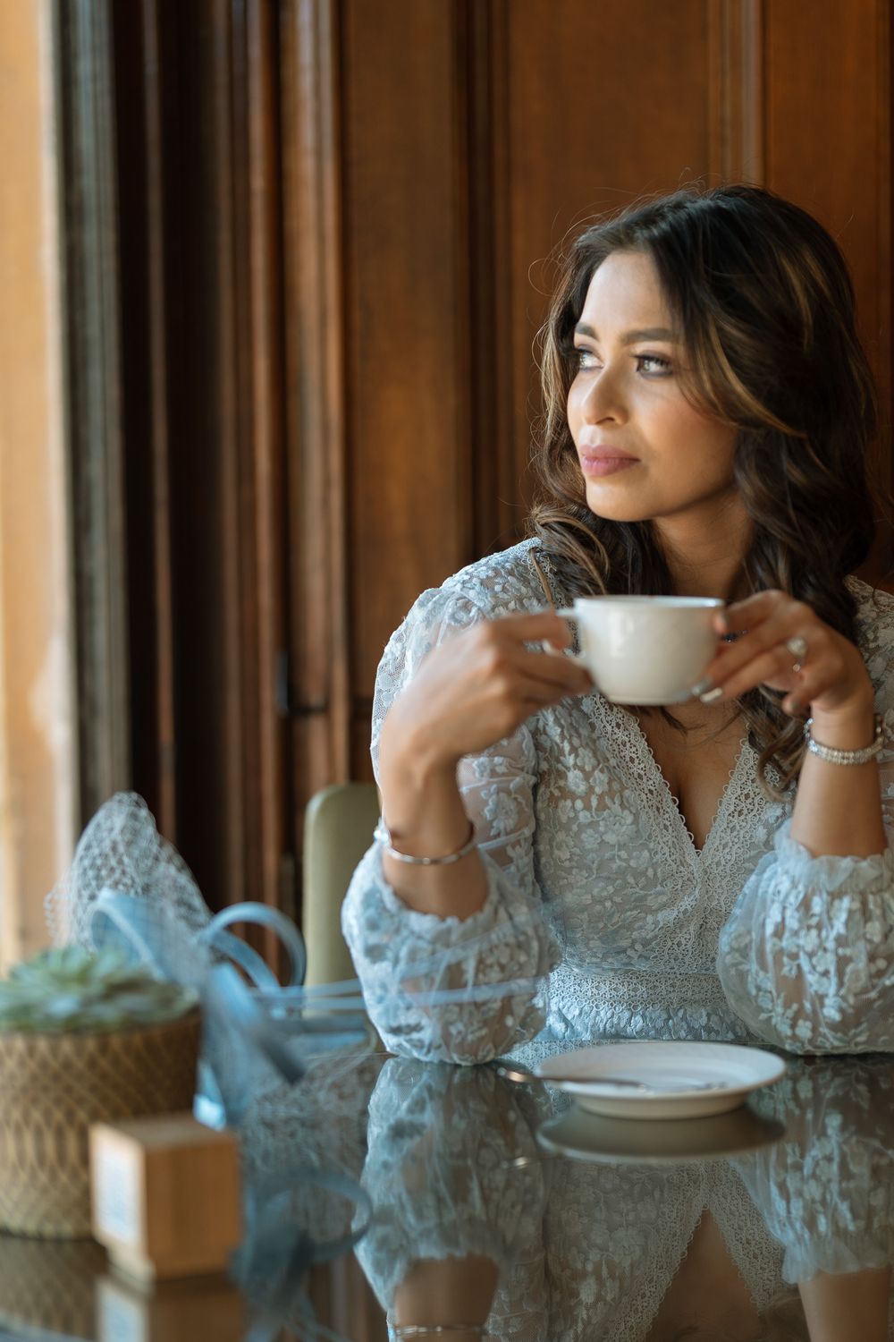 Elegant woman in white lace dress holding a coffee cup, gazing out a window in a sophisticated interior setting.