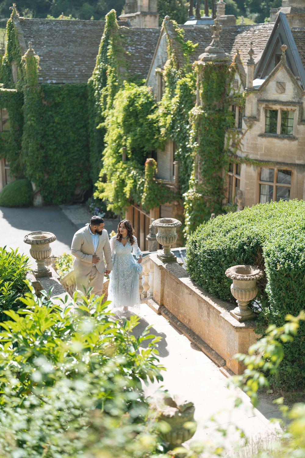 Couple walking through ivy-covered English manor gardens with stone urns and manicured hedges on a sunny day.