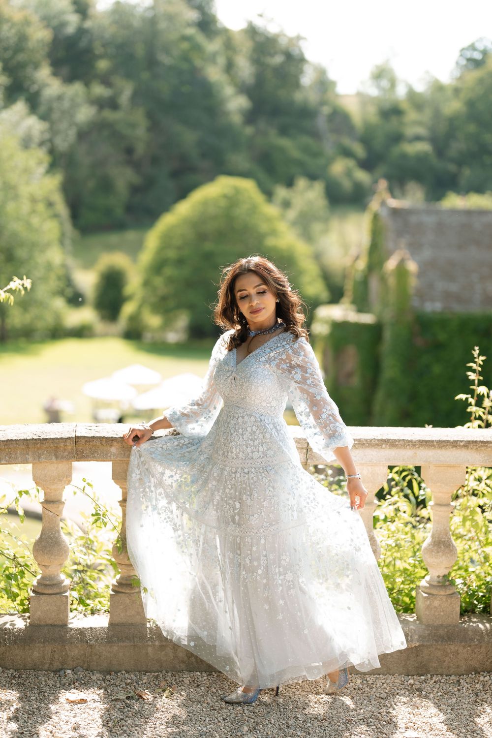 Beautiful woman in flowing white floral lace gown posing on stone balustrade surrounded by lush garden greenery.
