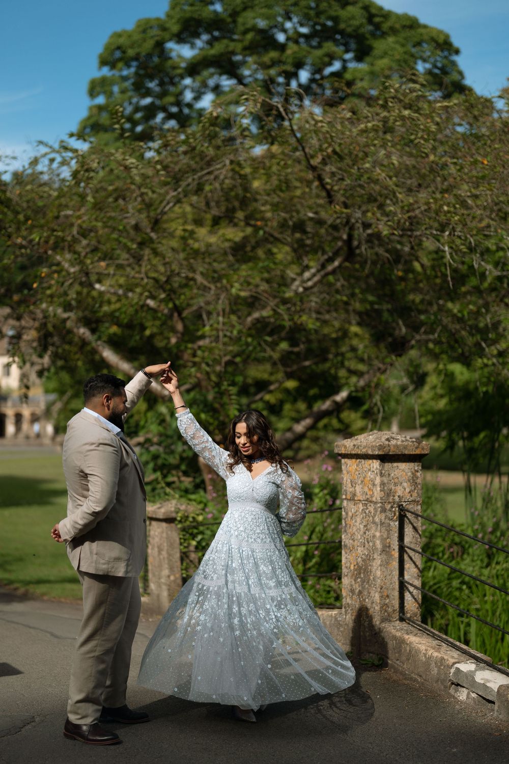 Couple dancing outdoors in a lush green park, bride in a flowing blue lace gown, groom in a beige suit.