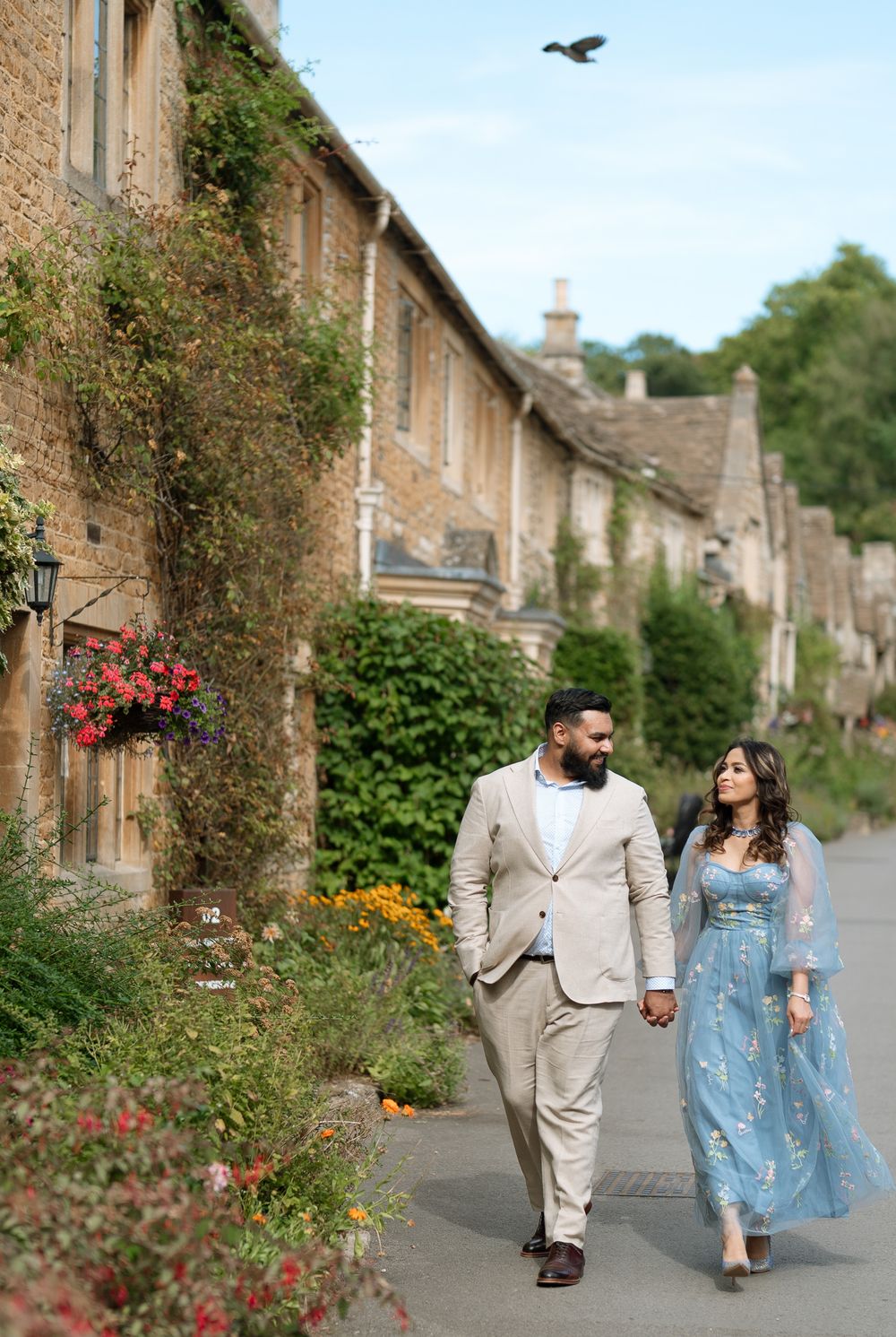 Couple walking hand-in-hand past flower-adorned Cotswolds stone cottages, man in beige suit, woman in blue floral gown.