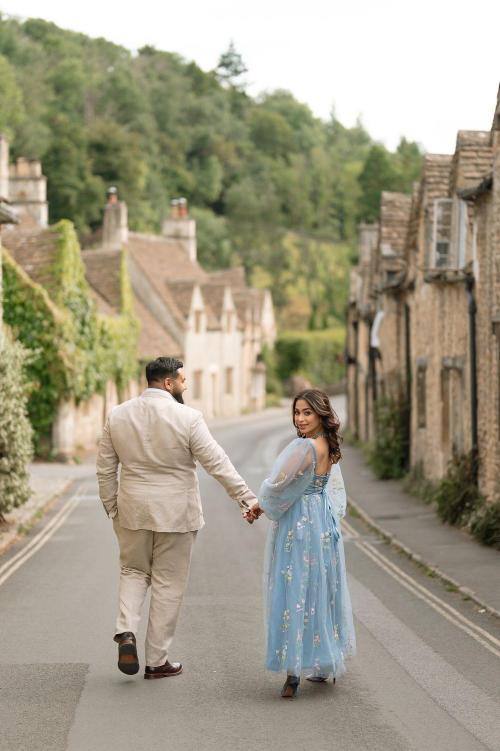 Couple holding hands walking down a charming Cotswolds village street, woman in blue floral gown glancing back.