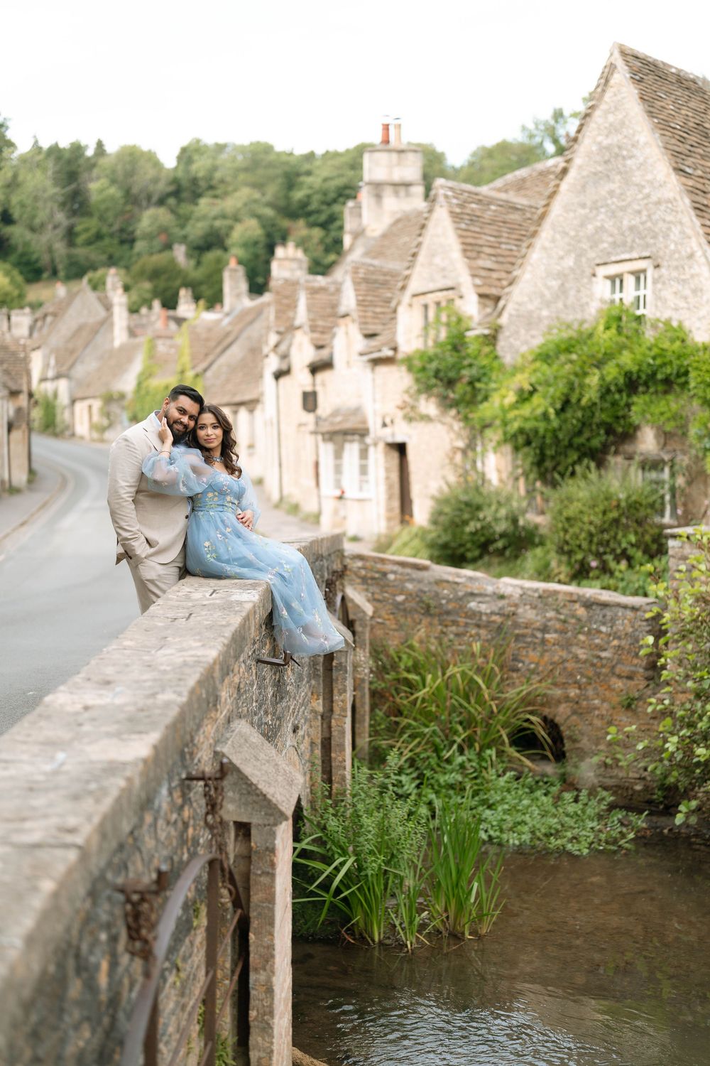 Couple sitting on a stone bridge in Castle Combe Cotswolds village, woman in blue dress, man in beige suit.