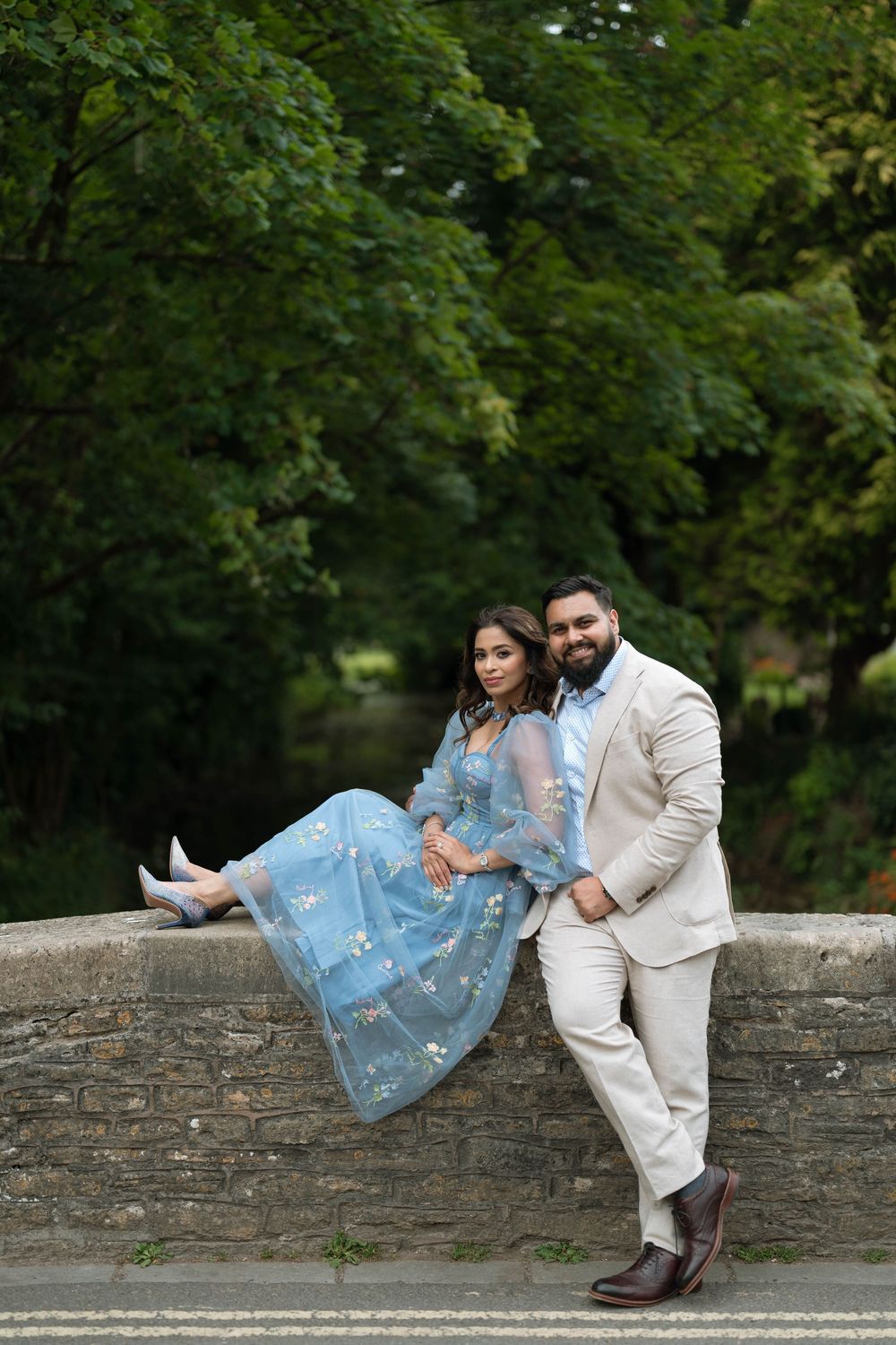 Couple posing on stone bridge, woman in blue floral dress seated, man in beige suit standing, lush green trees background.