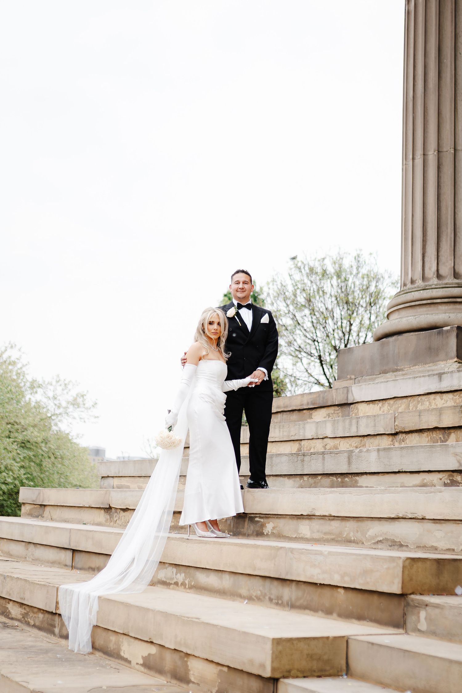 Black and white photo of a bride in white gown and gloves holding a bouquet, standing near stone steps.