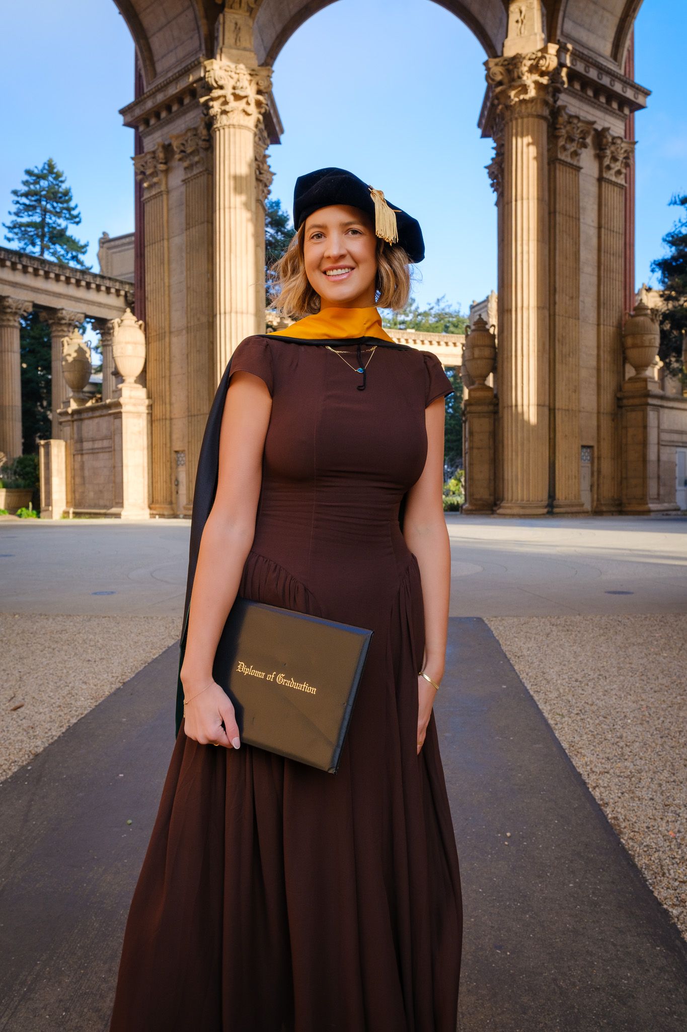 Smiling graduate in brown gown and black cap holds diploma at Palace of Fine Arts, San Francisco.