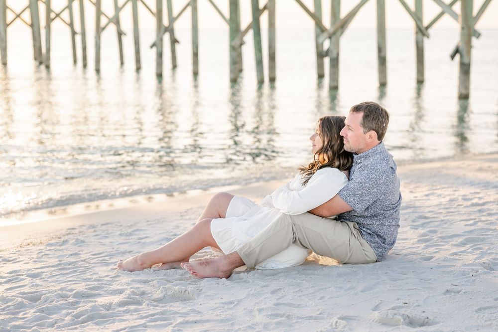 Couple sitting together on sandy beach near pier at sunset, sharing a romantic embrace in casual summer attire.