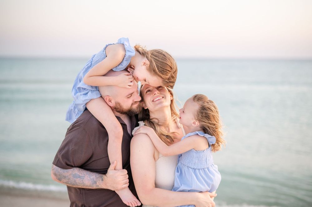 Family of four sharing joyful beach moment, parents holding two young girls in blue dresses at sunset.