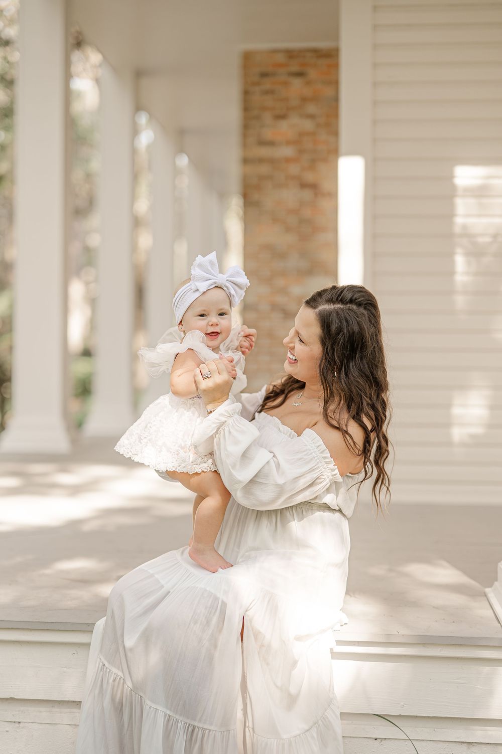 A mother in a white off-shoulder dress holds her smiling baby girl in a white bow on a sunlit porch.