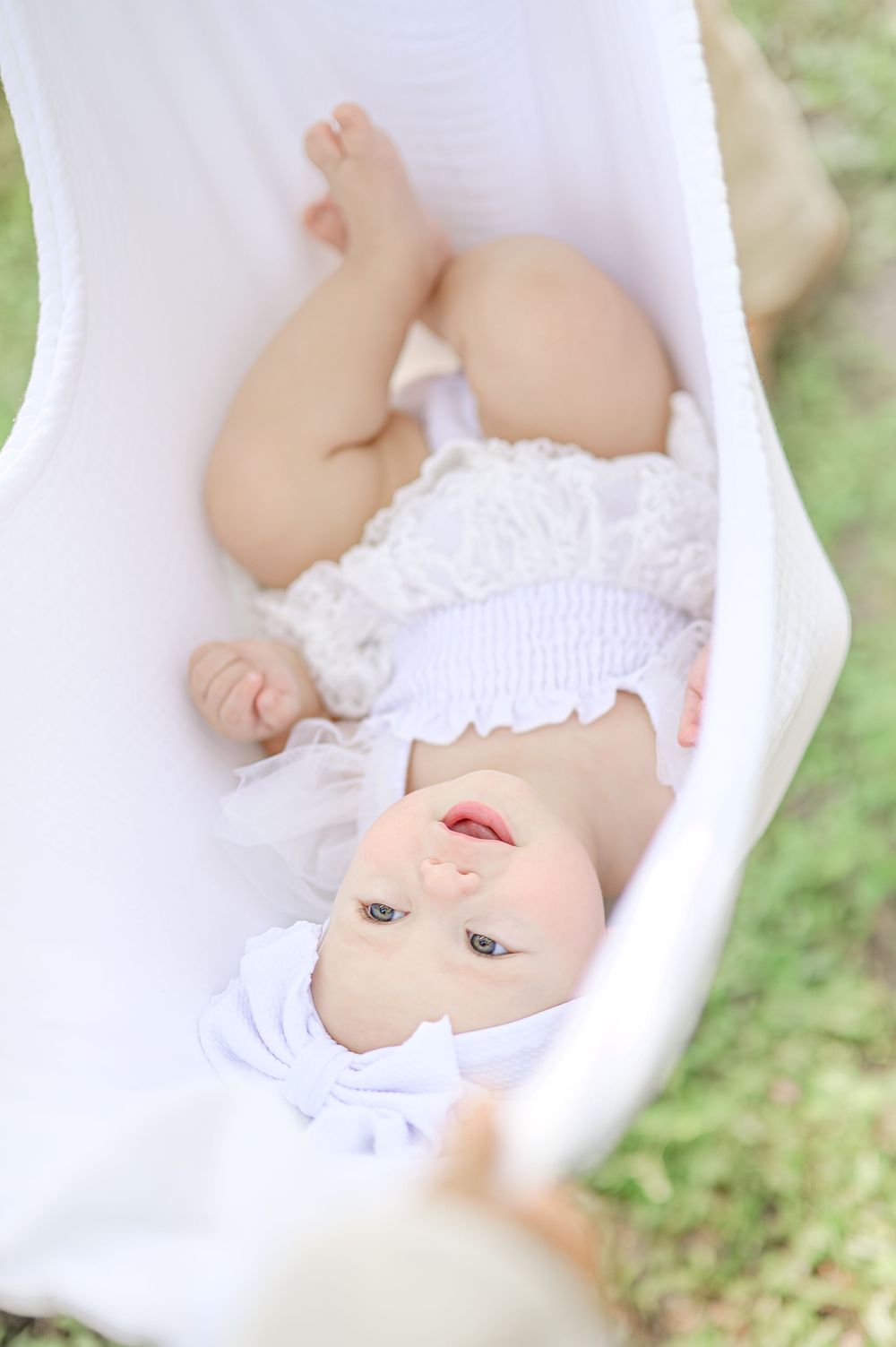 A smiling baby with blue eyes lies in a white bassinet outdoors, wearing a white ruffled outfit on green grass.