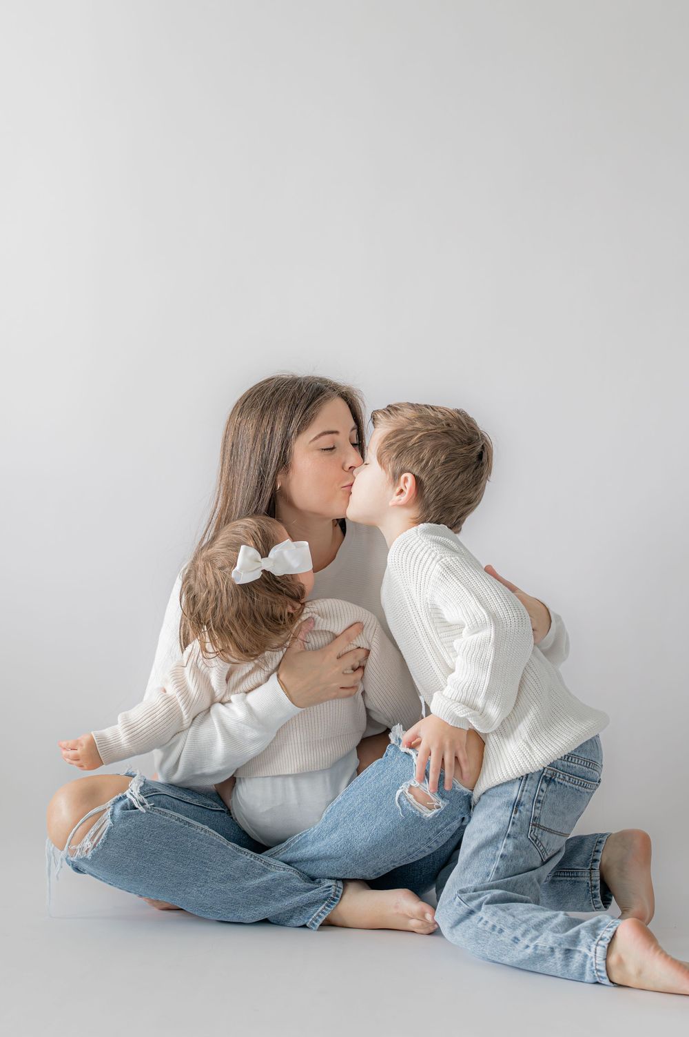 A mother sits cross-legged kissing her son while holding her young daughter, all wearing white tops and jeans.