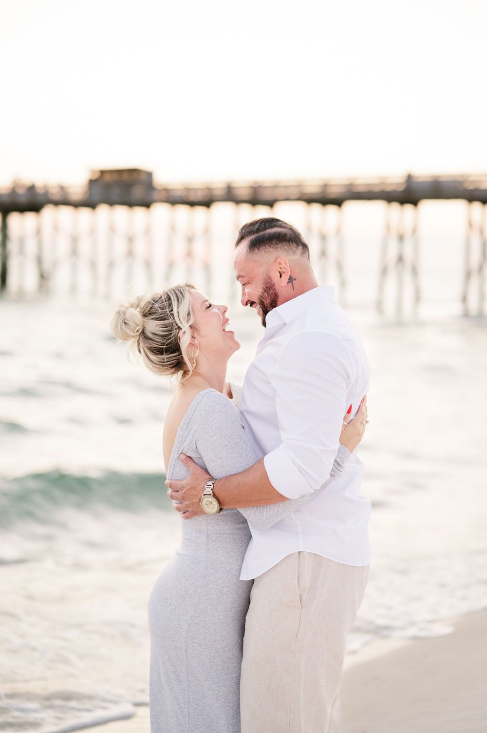 A couple embraces and laughs together on a beach near a pier during a romantic golden hour photo session.