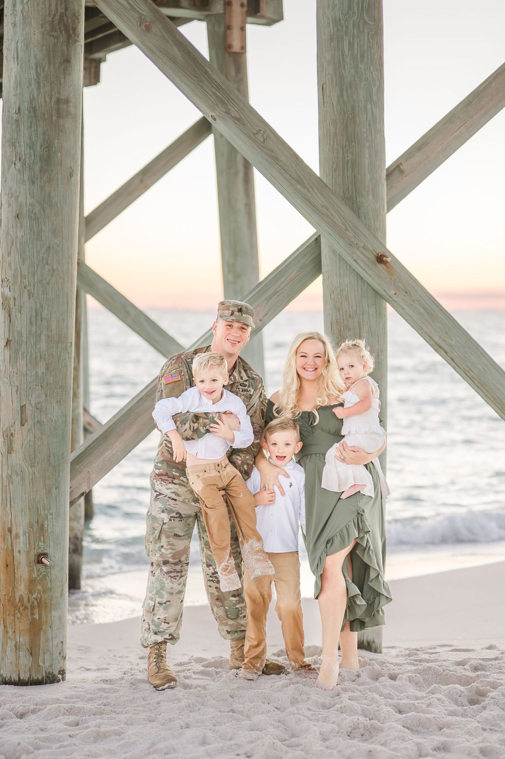 Military family poses under a beach pier at sunset, father in camo uniform holding child, mother in green dress holding baby.
