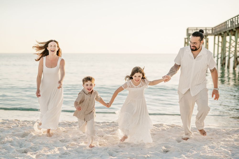 Family of four joyfully running on a sandy beach near a pier, dressed in white and beige outfits.