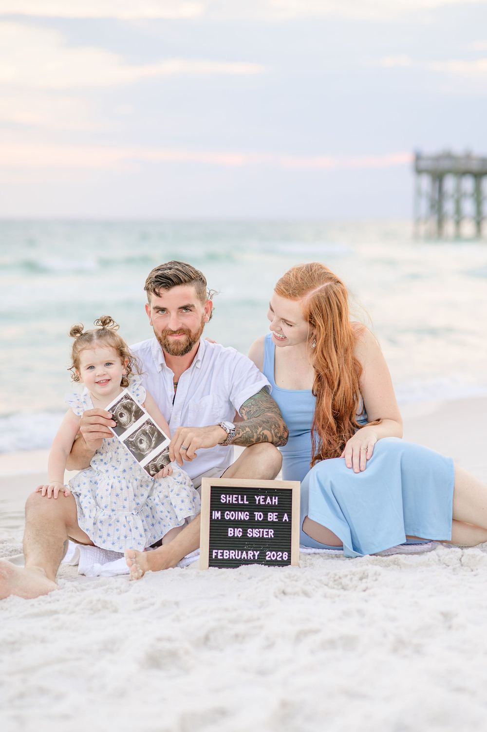 Family pregnancy announcement on beach with ultrasound photo and letter board reading big sister reveal February 2028.