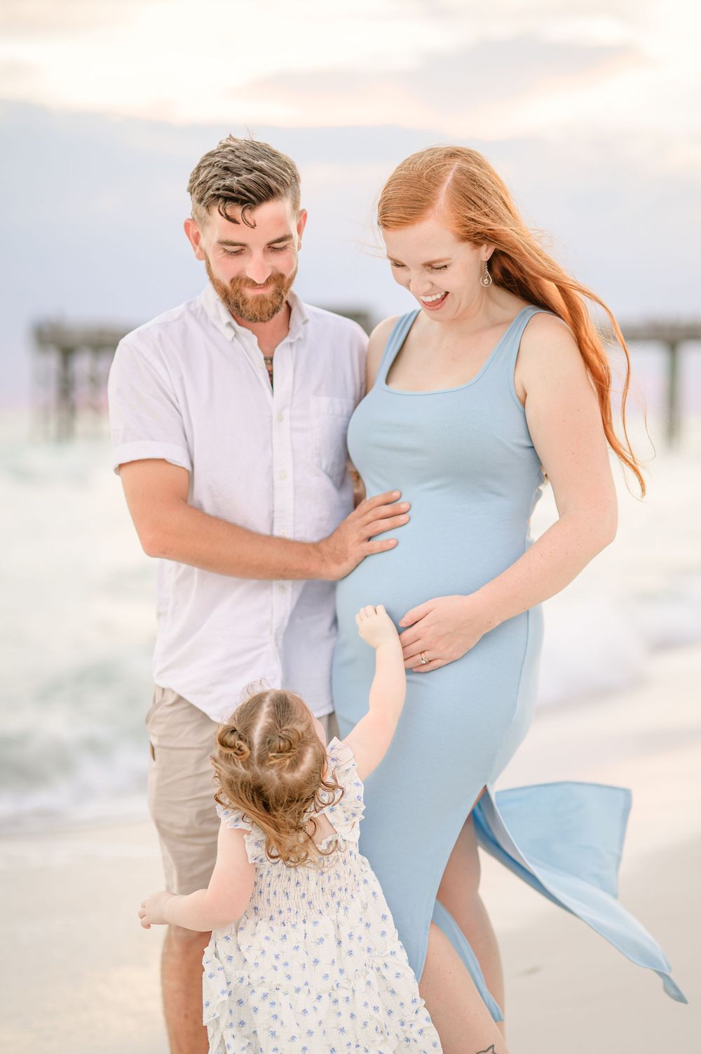 Pregnant woman in blue dress with husband and toddler daughter on beach at sunset.