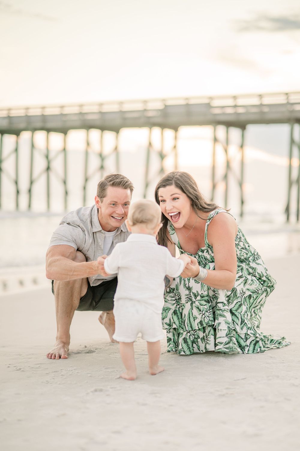 A joyful family of three enjoys a golden hour beach session near a pier, with parents cheering their baby's first steps.