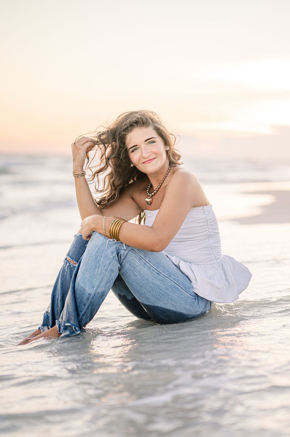 Young woman in white top and jeans sitting on a beach at sunset, smiling with waves gently washing around her.