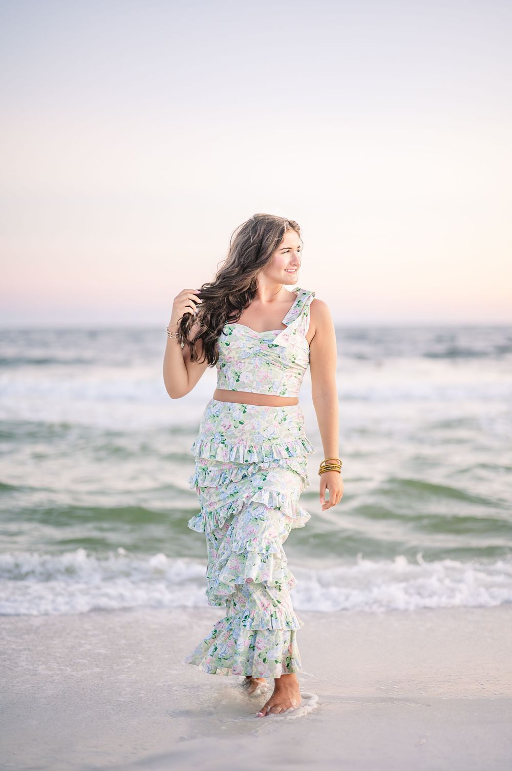 Young woman in floral two-piece outfit standing at the ocean's edge during sunset at the beach.