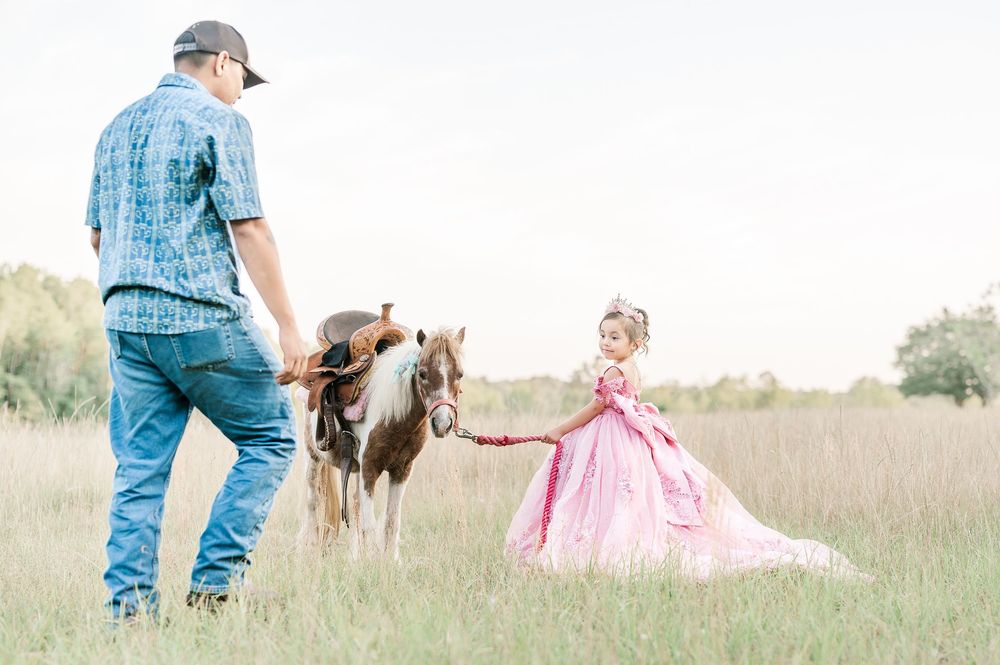 A man and young girl in a pink gown walk a pinto pony through a grassy field.