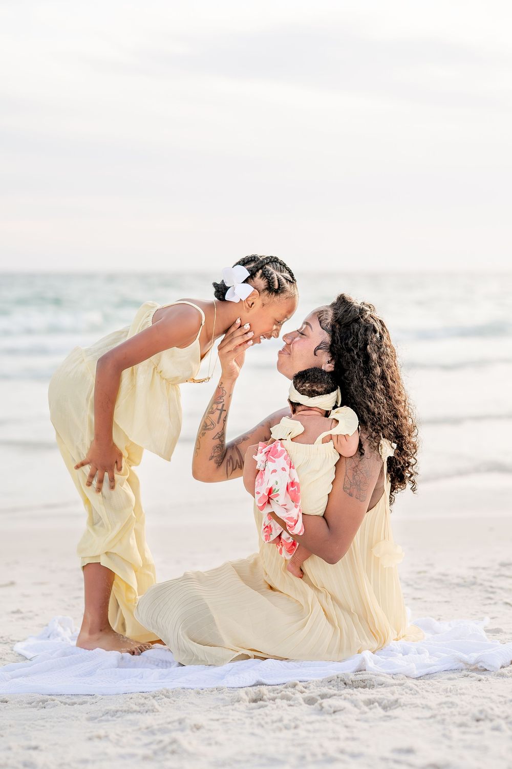 A mother and daughter in matching yellow dresses share a tender moment on a sandy beach near the ocean.