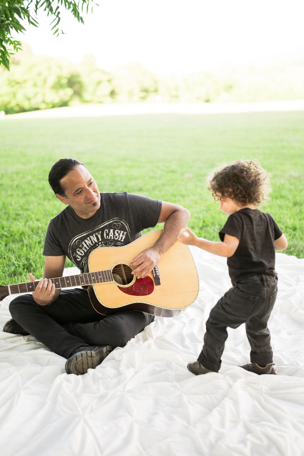 Hispanic Father in Johnny Cash shirt plays acoustic guitar for curly-haired toddler son on white blanket in sunny park. Family Photographer.