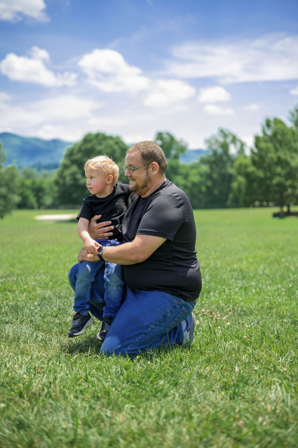 A father kneeling on green grass holds his young blonde son outdoors, with trees and mountains in the background. Family Photographer.