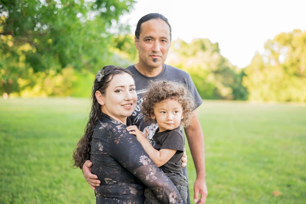 A happy hispanic family of three poses outdoors in a park, with parents holding their young curly-haired child. Family Photographer.
