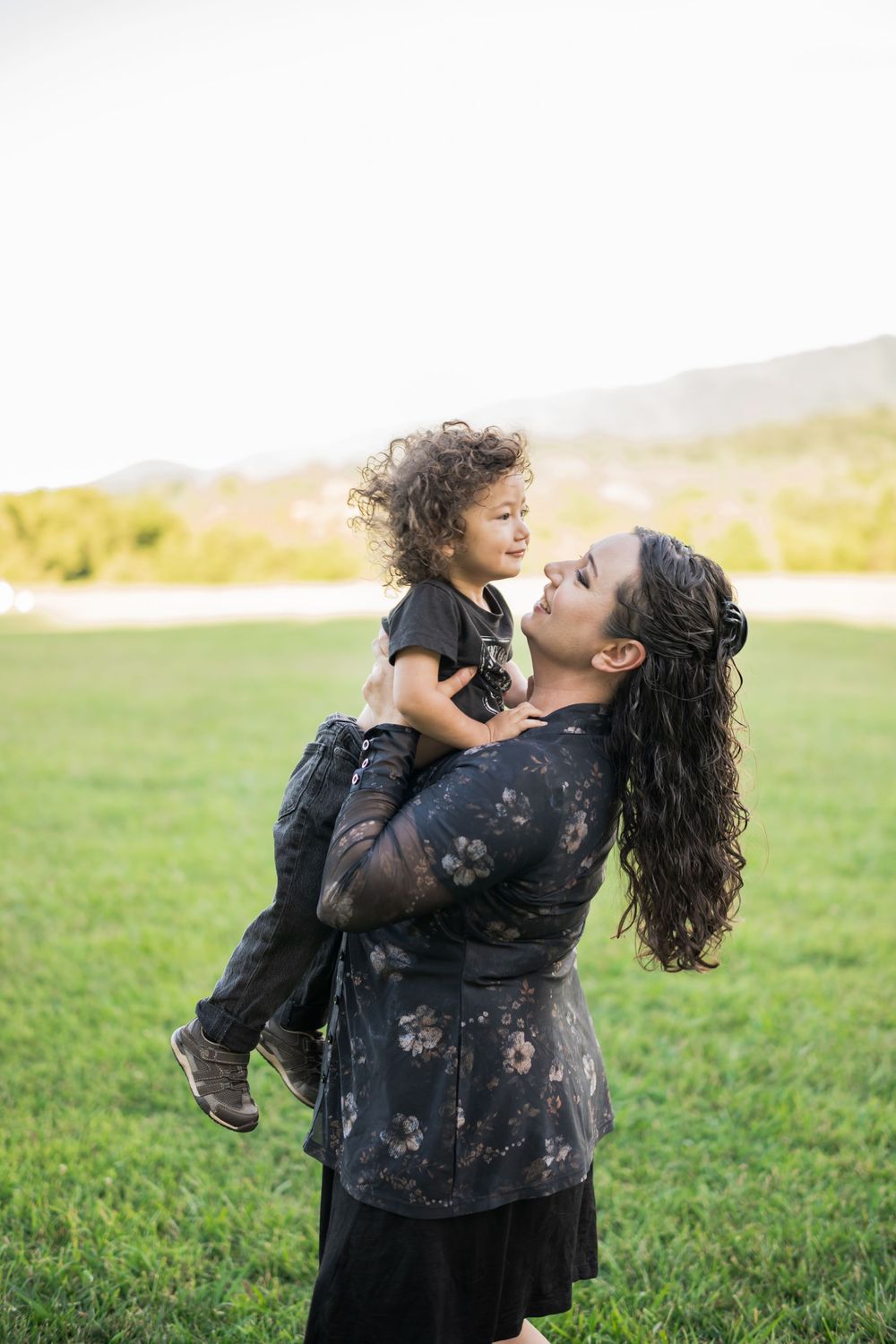 A hispanic mother holds her curly-haired toddler in a green field, both smiling and dressed in black outfits. Family Photographer.