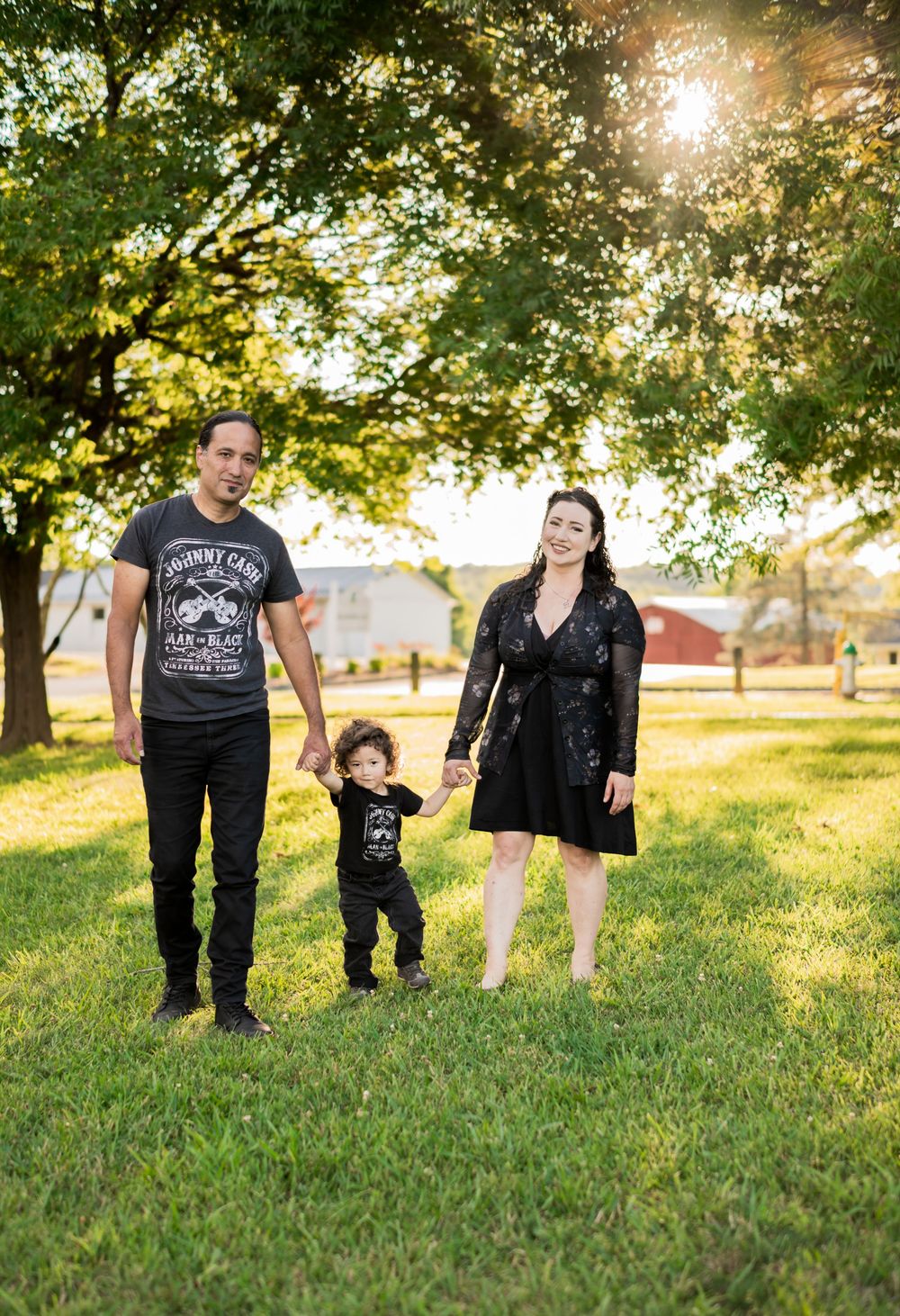 Hispanic Family walking hand-in-hand on grass under sunlit trees, parents in black with young toddler between them. Family Photograrapher.