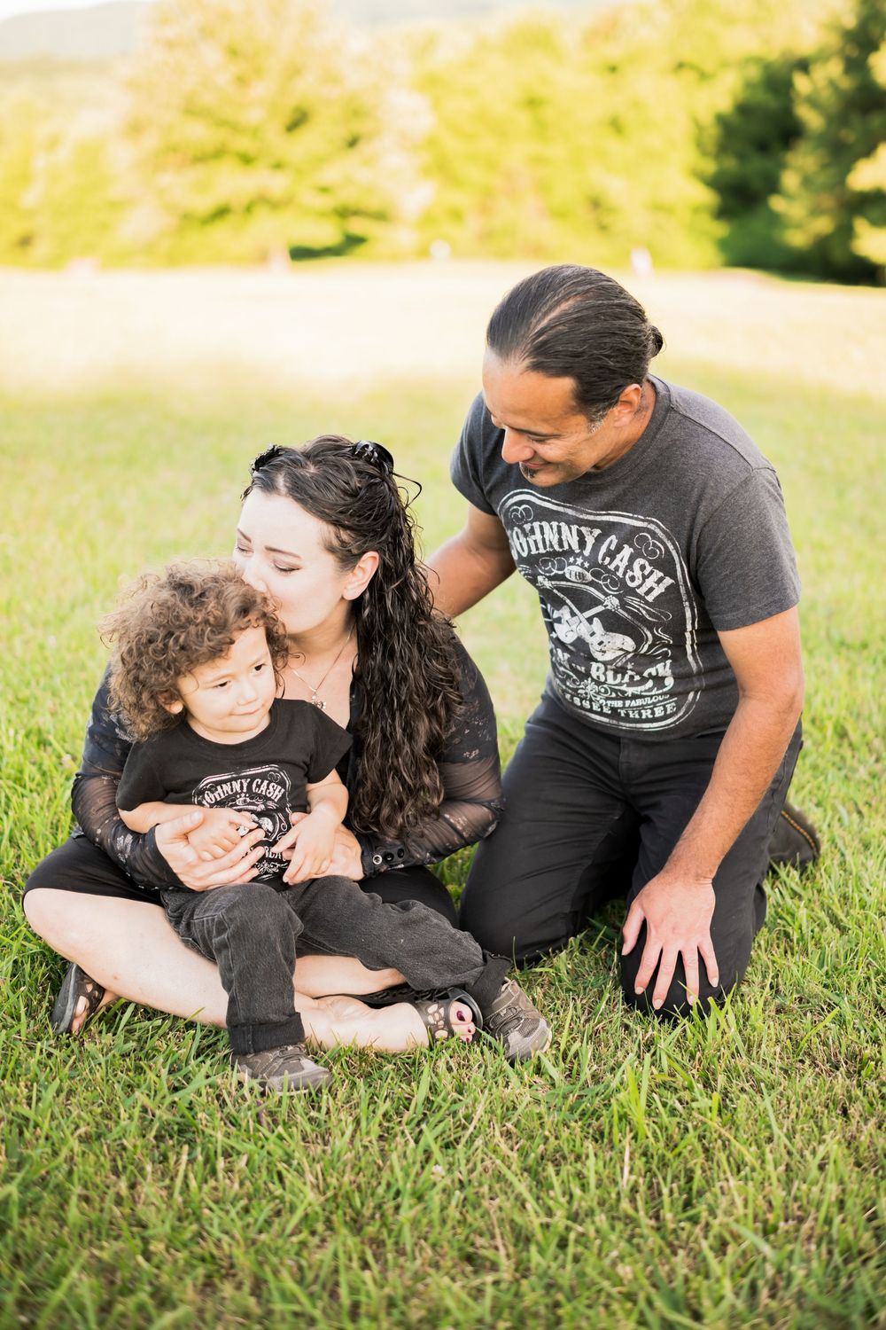 Hispanic family of three sitting on grass outdoors, parents wearing Johnny Cash shirts, with their curly-haired toddler. Family Photographer