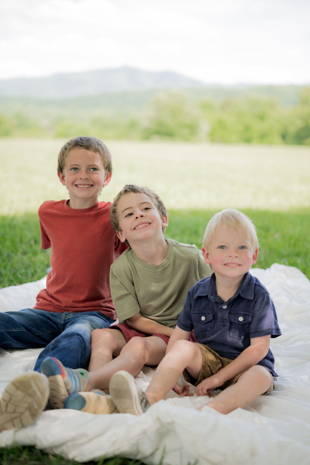 Three smiling boys sitting on a white blanket outdoors with green fields and mountains in the background. Family Photographer.