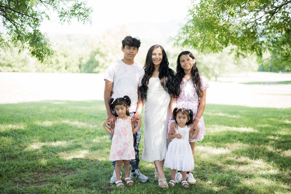 Family of five posing outdoors on green grass, mother in white dress with four children, surrounded by trees. Family/Maternity Photography.