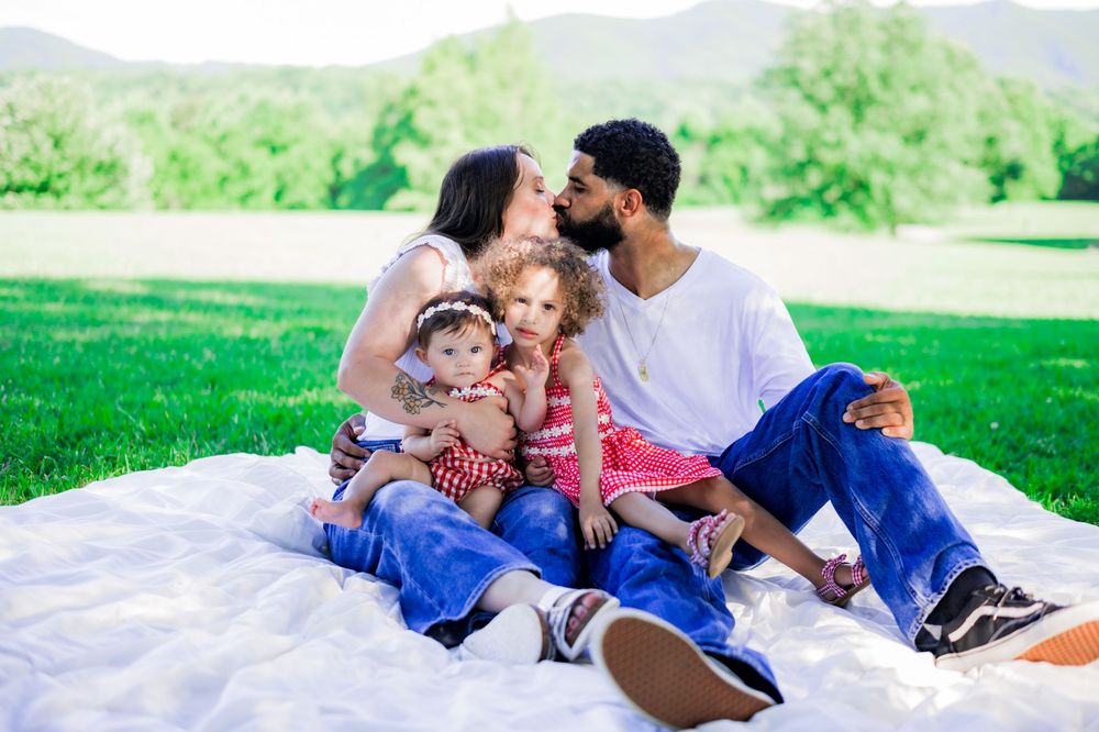 Happy family of four sitting on a blanket in a green park, parents kissing their young children outdoors. Family Photographer.