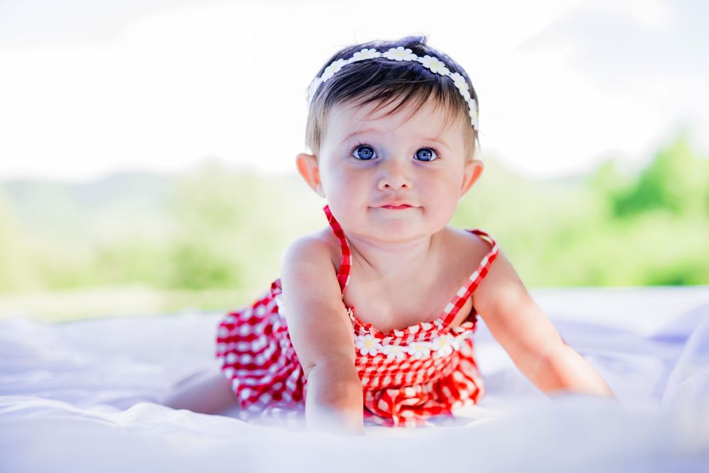 Adorable baby girl with blue eyes wearing a red dress and headband, crawling on a white blanket outdoors. Family Photographer.