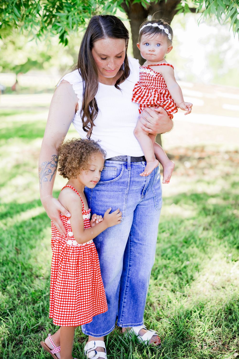 A mother in jeans holds a baby in red gingham while her curly-haired toddler hugs her leg outdoors. Family Photographer.