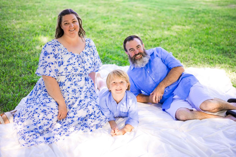 Family of three smiling on a white blanket in green grass, wearing coordinating blue and white outfits. Family Photographer.